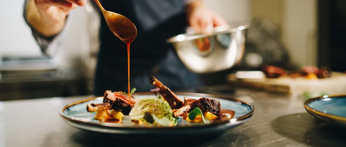 Chef drizzling red sauce over a plated dish of lamb chops with microgreens.