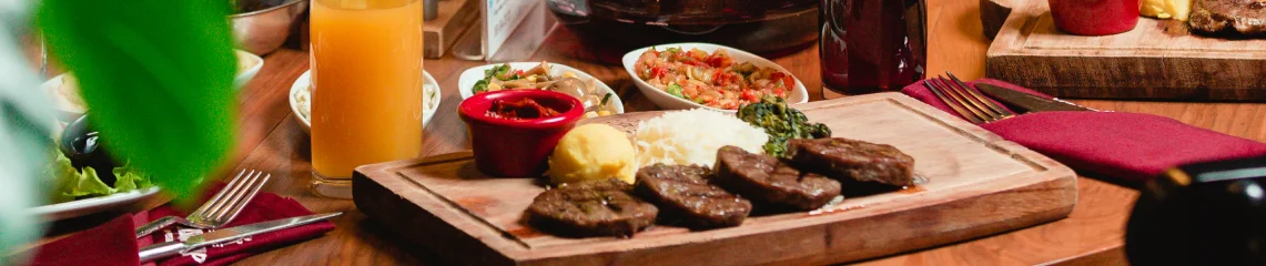 Wooden serving board with grilled steak medallions, rice, and sides alongside orange juice on a dining table